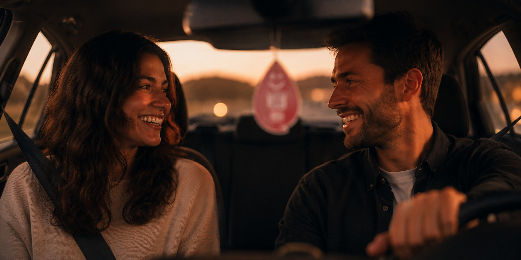 Paar lacht im Auto bei warmer Abendstimmung mit Duftanhänger am Rückspiegel